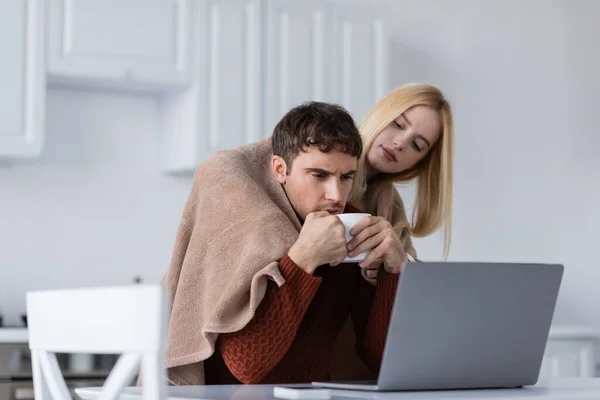 Donna bionda che tiene la coperta vicino al fidanzato con tazza che lavora a distanza da casa — Foto stock