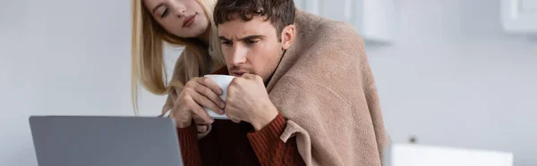 Donna bionda che tiene la coperta vicino al fidanzato con tazza che lavora a distanza da casa, banner — Foto stock