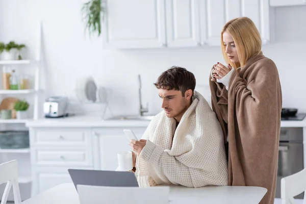 Mulher loira coberta de cobertor perto do namorado com smartphone e xícara — Fotografia de Stock