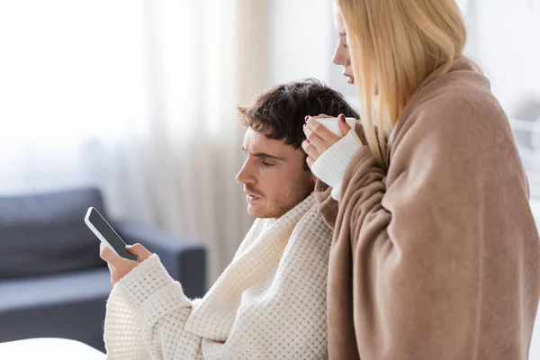 Femme blonde couverte d'une couverture tenant la tasse près du petit ami en utilisant un smartphone près d'un ordinateur portable — Photo de stock