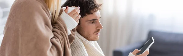 Mujer cubierta de manta que sostiene la taza cerca del novio usando el teléfono inteligente con la pantalla en blanco, bandera — Stock Photo