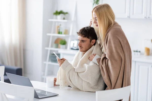 Blonde woman covered in blanket standing near boyfriend with smartphone and cup of tea — Stock Photo