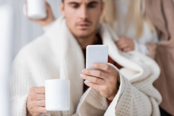 Homme flou en utilisant smartphone et tenant tasse de thé près de petite amie — Photo de stock
