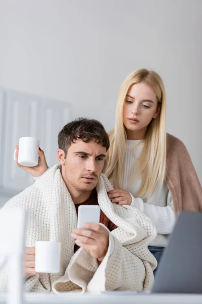 Young woman covered in blanket standing near boyfriend with smartphone and cup of tea — Stock Photo