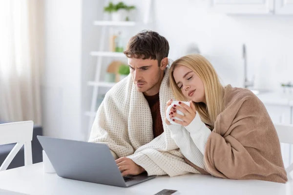 Mujer rubia joven cubierta de manta apoyada en el hombro del novio usando el ordenador portátil en casa - foto de stock