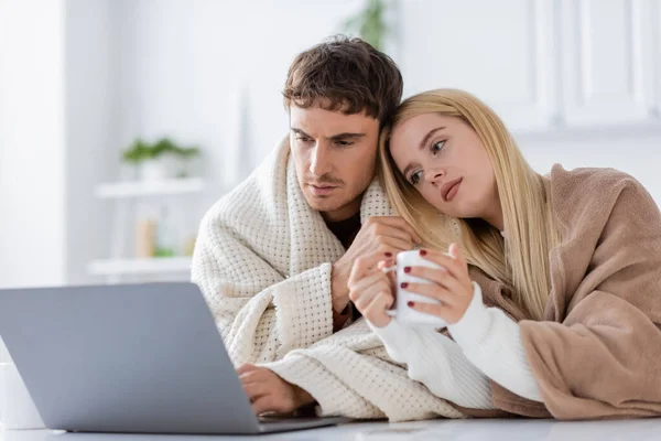 Young blonde woman covered in blanket leaning on shoulder of boyfriend using laptop and working from home — Stock Photo