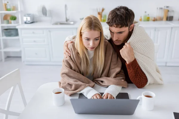 Vista de ángulo alto de la mujer rubia utilizando el ordenador portátil cerca de novio y tazas en la mesa - foto de stock