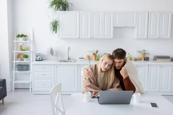 Young blonde woman covered in blanket leaning on shoulder of boyfriend near gadgets and cups on table — Stock Photo