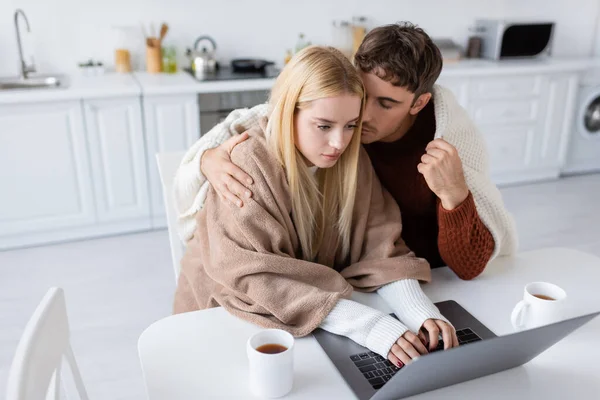 Hochwinkelaufnahme der blonden Frau mit Laptop, während ihr Freund sie in der Nähe von Tassen auf dem Tisch umarmt — Stockfoto