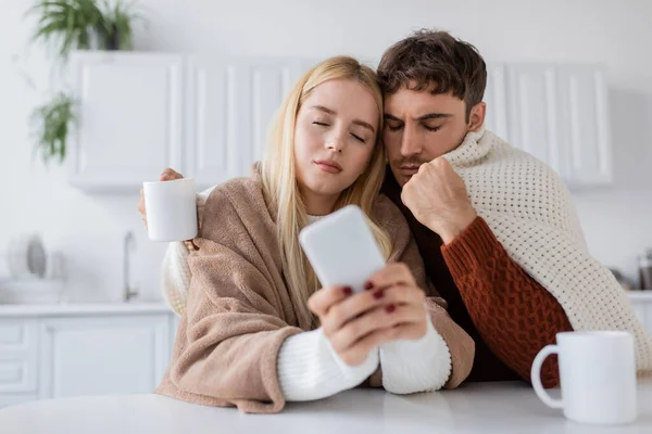 Rubia joven utilizando el teléfono móvil cerca de novio y taza en la mesa - foto de stock