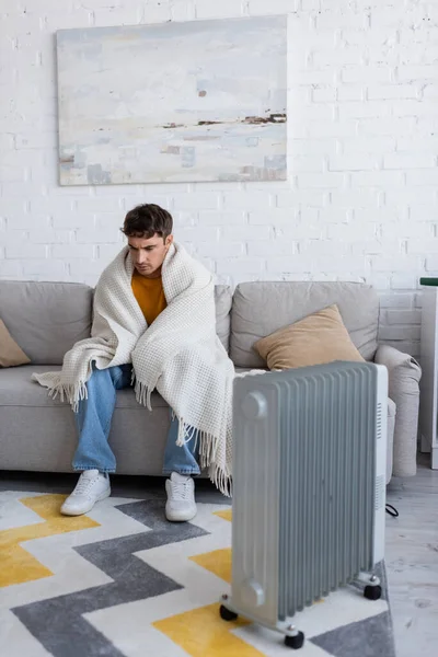Comprimento total do jovem coberto de cobertor sentado no sofá perto do aquecedor do radiador no inverno — Fotografia de Stock