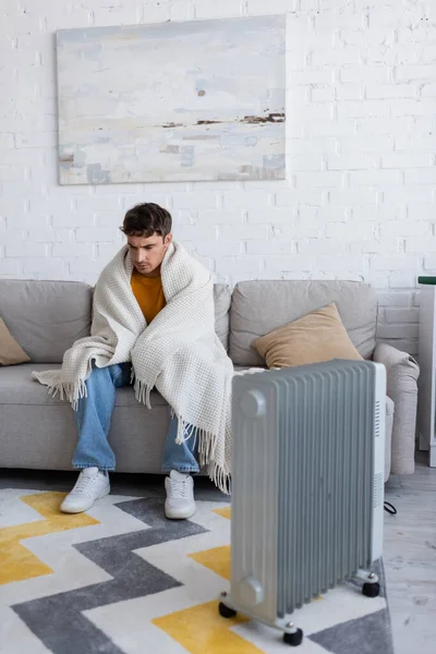 Junge Mann in Decke gehüllt sitzt im Winter auf Sofa neben Heizkörper — Stockfoto