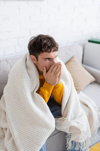 Jeune homme couvert de couverture assis sur le canapé et se réchauffant les mains dans le salon — Photo de stock