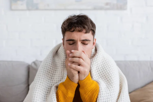 Молодой человек с закрытыми глазами сидит покрытый одеялом и греет руки в гостиной — стоковое фото