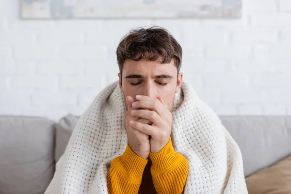 Jeune homme les yeux fermés assis couvert de couverture et les mains chaudes dans le salon — Photo de stock