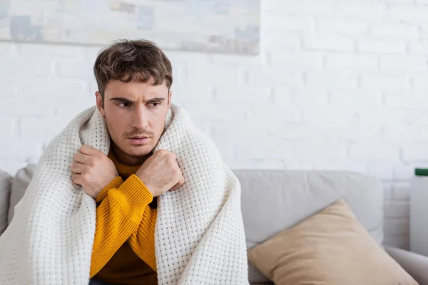 Junger Mann in Decke gehüllt auf Sofa sitzend, während er sich im Wohnzimmer warm macht — Stockfoto