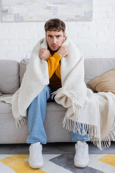Розчарований молодий чоловік, покритий ковдрою, сидить на дивані, зігріваючись у вітальні — стокове фото