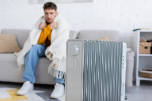 Radiador calentador cerca de hombre borroso cubierto de manta sentado en el sofá en la sala de estar - foto de stock