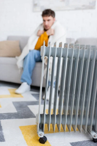 Обогреватель радиатора рядом с размытым молодым человеком, покрытым одеялом, сидящим на диване в гостиной — стоковое фото