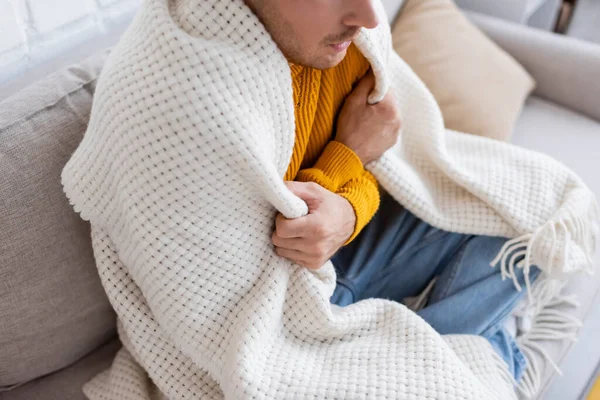 Vista de ángulo alto de hombre joven cubierto de manta sentado en el sofá y calentándose en la sala de estar - foto de stock