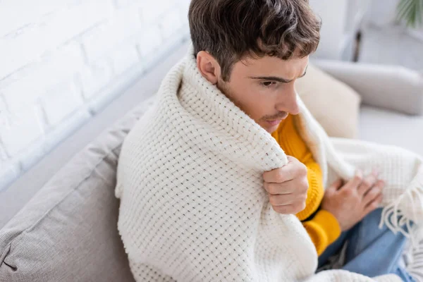 Высокий угол зрения молодого человека, покрытого одеялом, сидящего на диване и согревающегося дома — стоковое фото