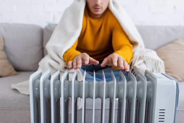 Ausgeschnittener Blick auf einen jungen Mann, der in eine Decke gehüllt auf dem Sofa sitzt und sich im Winter in der Nähe der Heizkörperheizung aufwärmt — Stockfoto