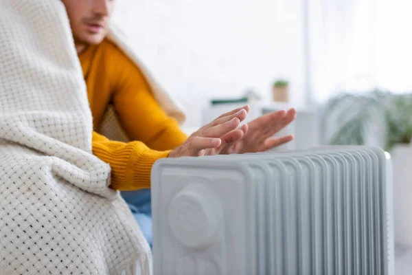 Partial view of young man covered in blanket sitting on sofa and warming up near radiator heater — Stock Photo