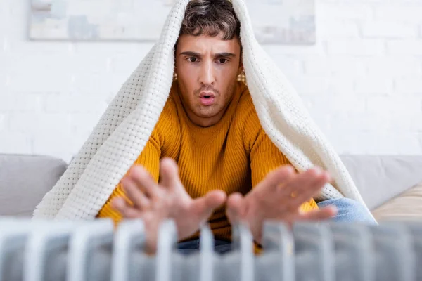 Emotional young man covered in blanket warming up hands near radiator heater — Stock Photo