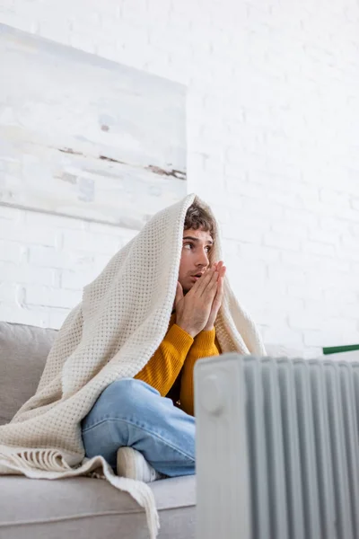 Young man covered in blanket sitting on sofa and warming up hands near radiator heater — Stock Photo