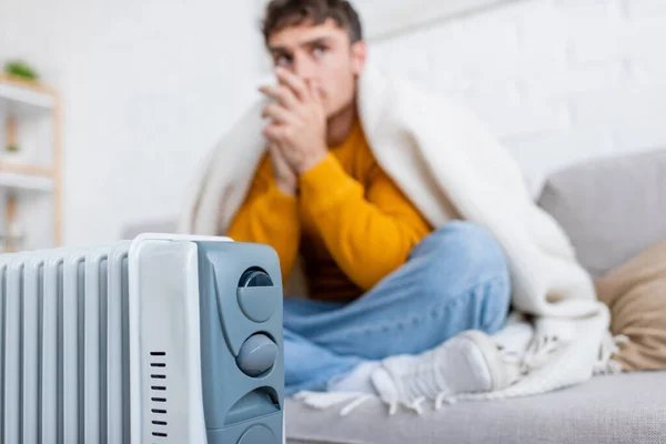 Moderno calentador de radiador cerca de hombre borroso cubierto de manta sentado en el sofá - foto de stock