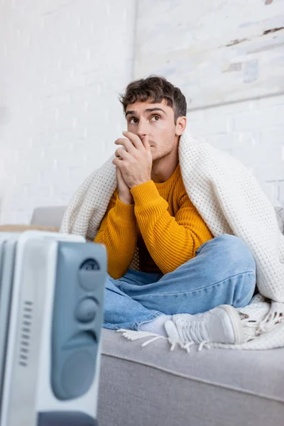 Jeune homme couvert de couverture assis sur le canapé près du chauffage flou en hiver — Photo de stock