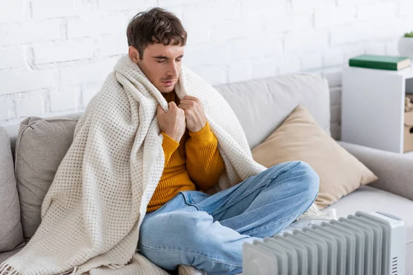 Молодой человек с закрытыми глазами держит одеяло, сидя на диване рядом с обогревателем радиатора зимой — стоковое фото