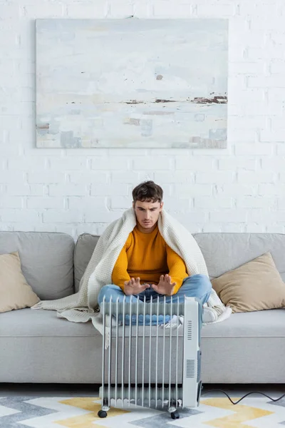 Hombre joven cubierto de manta sentado en el sofá y las manos de calentamiento cerca del radiador moderno - foto de stock