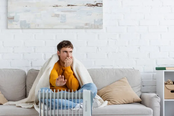 Junger Mann in Decke gehüllt auf Couch sitzend und wärmende Hände neben modernem Heizkörper — Stockfoto