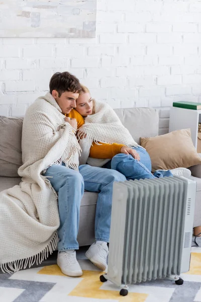 Молода пара вкрита ковдрою, сидить на дивані і зігрівається біля сучасного радіатора — стокове фото