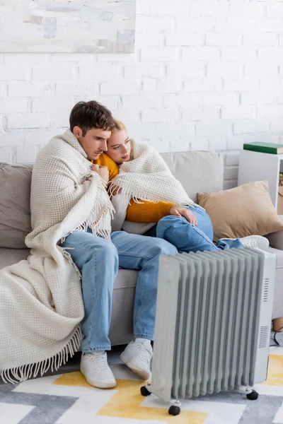 Jeune couple couvert de couverture assis sur le canapé et se réchauffant près du radiateur moderne — Photo de stock