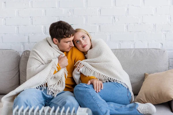 Jeune couple couvert de couverture assis sur le canapé et se réchauffant près du radiateur moderne — Photo de stock