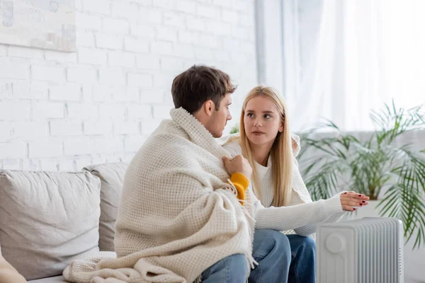 Jeune couple couvert de couverture assis sur le canapé et se réchauffant près du radiateur à la maison — Photo de stock