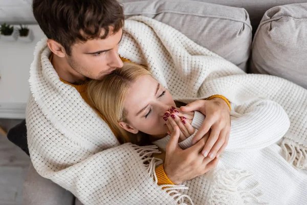 Vista superior do homem coberto de cobertor beijando cabeça de mulher loira em suéter branco aquecendo as mãos — Fotografia de Stock