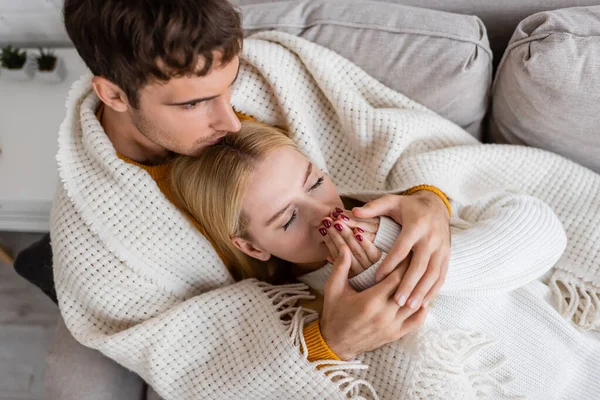 Vista dall'alto dell'uomo coperto di coperta baciare la testa della donna bionda in maglione bianco riscaldando le mani — Foto stock