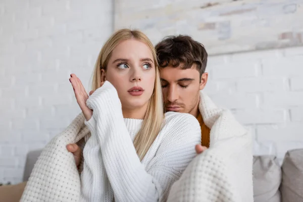 Homme attentionné couvrant petite amie blonde en pull blanc avec couverture à la maison — Photo de stock