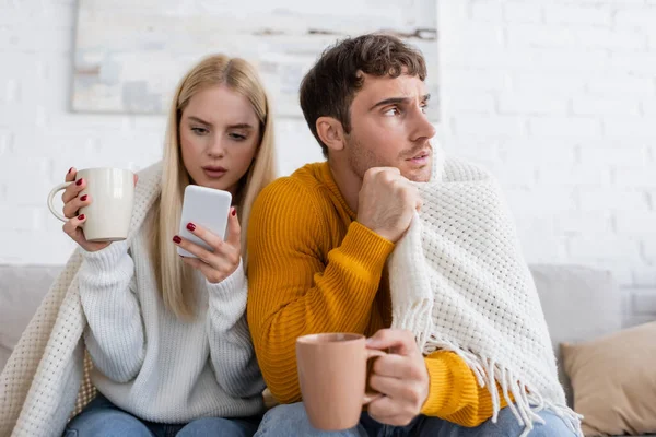 Mujer joven sosteniendo la taza de té y el uso de teléfono inteligente cerca de novio cubierto con manta - foto de stock