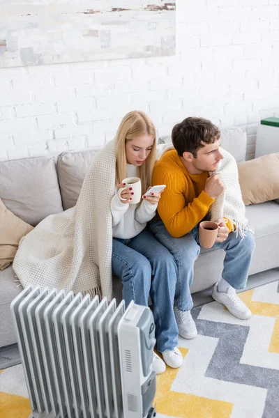 Jeune femme tenant une tasse de thé et utilisant un smartphone près du petit ami recouvert d'une couverture assise près du radiateur — Photo de stock