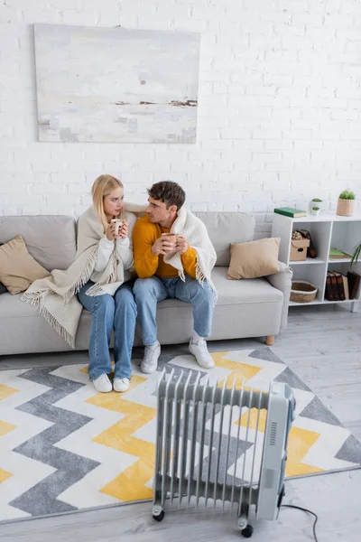 Pareja joven cubierta de manta sentada con tazas de té cerca del radiador - foto de stock