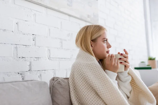 Junge Frau im Pullover sitzt in Decke gehüllt mit Tasse Tee im Wohnzimmer — Stockfoto