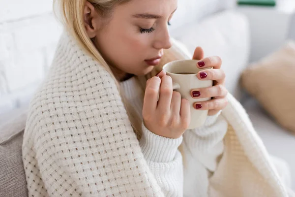 Young blonde woman in white sweater sitting covered in blanket with cup of tea — Stock Photo