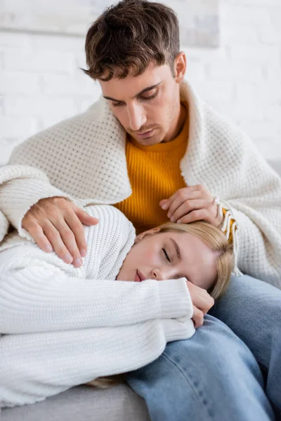 Mujer joven cubierta de manta durmiendo en las piernas de novio cariñoso en jeans sentado en el sofá - foto de stock