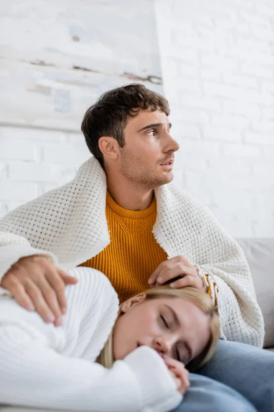 Donna bionda coperta di coperta che dorme sulle gambe del fidanzato in jeans seduta sul divano — Foto stock