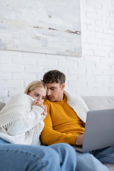 Испуганная молодая пара, покрытая одеялом, смотрит страшный фильм на ноутбуке в гостиной — стоковое фото