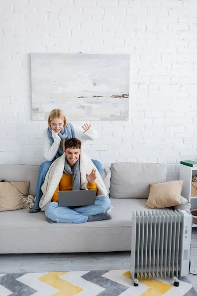 Alegre pareja en bufandas y suéteres agitando las manos durante la videollamada en el ordenador portátil - foto de stock